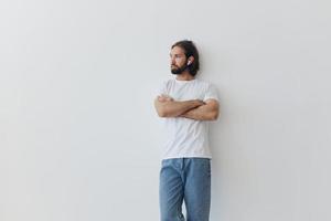 une homme avec une barbe et longue cheveux dans une blanc T-shirt et bleu jeans des stands contre une blanc mur, penché contre il et écoute à la musique avec sans fil blanc écouteurs, regarder pensivement photo