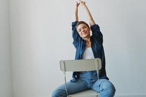 une Jeune femme séance dans une chaise à Accueil souriant avec les dents avec une court la Coupe de cheveux dans jeans et une denim chemise sur une blanc Contexte. fille Naturel pose avec non filtres photo