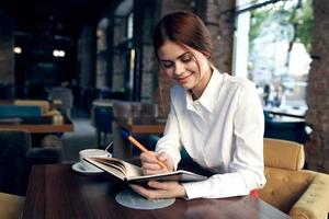 une femme dans une chemise avec une carnet et une stylo dans sa main est assis à une table dans une restaurant sur un tapissé chaise photo