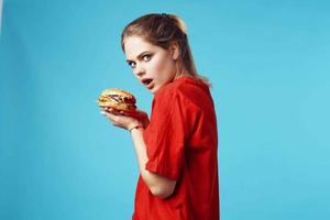 de bonne humeur femme dans rouge T-shirt Hamburger dans mains vite nourriture bleu Contexte photo