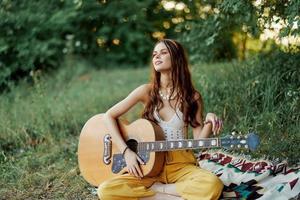 fille hippie femme en jouant guitare dans respectueux de la nature vêtements séance sur le sol à l'extérieur dans la nature dans le tomber en train de regarder le le coucher du soleil photo