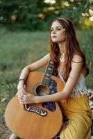 hippie femme éco amical en jouant guitare sourit et en chantant Chansons dans la nature séance sur une plaid par le Lac dans le soir dans le le coucher du soleil lumière du soleil. une mode de vie dans harmonie avec le corps et la nature photo