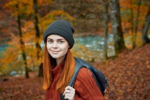 énergique femme touristique avec une sac à dos dans une rouge chandail et casquettes sont repos dans le l'automne forêt dans le parc photo