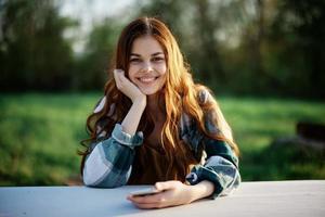 fille avec téléphone dans main séance dans la nature dans le parc à le table souriant magnifiquement et à la recherche à le caméra avec sa rouge cheveux allumé par le le coucher du soleil lumière du soleil de été photo