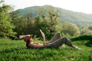 content femme blogueur mensonge sur le herbe dans le parc et souriant avec sa téléphone dans sa mains contre le toile de fond de une été Naturel paysage avec lumière du soleil photo