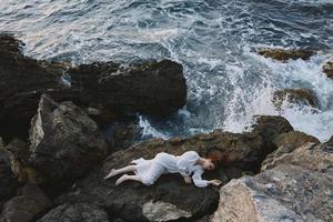 pieds nus femme dans longue blanc robe humide cheveux mensonge sur une rocheux falaise inchangé photo