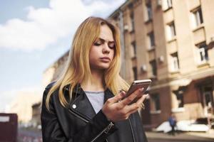 tondu vue de blond femme avec mobile téléphone sur le rue près le bâtiment photo