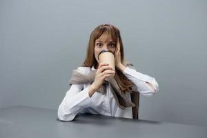 excité Jeune blond étudiant Dame pigiste dans chaud chandail boisson à emporter café Regardez à caméra séance à lieu de travail dans gris moderne Accueil bureau. café Pause amoureux concept. copie espace. cool offre photo