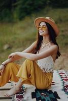une content fille séance dans éco Vêtements dans la nature sur une plaid par le Lac portant une chapeau dans une hippie Regardez et souriant dans merveille. l'automne mode de vie photo