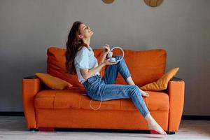 souriant femme écoute à la musique avec écouteurs sur le Orange canapé appartements photo
