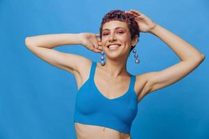 Jeune athlétique mode femme avec coloré cheveux et court la Coupe de cheveux posant et dansant dans bleu tenue de sport souriant et à la recherche à le caméra sur une bleu monochrome Contexte photo