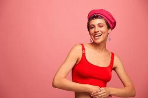 Jeune athlétique femme avec une court la Coupe de cheveux et violet cheveux dans une rouge Haut et une rose chapeau avec un athlétique figure sourit et grimaces à la recherche à le caméra sur une rose Contexte photo