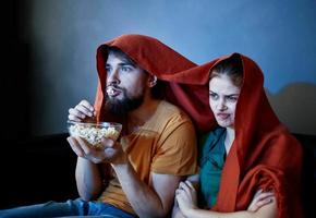 une homme avec une assiette de pop corn et un émotif femme en dessous de une rouge couverture sur le canapé photo