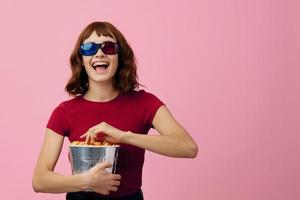 fou de joie content mignonne roux Dame dans rouge T-shirt 3d des lunettes avec pop corn regarder film posant isolé sur plus de rose studio Contexte. copie espace bannière. mode cinéma concept. divertissement offre photo