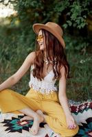 une Jeune hippie femme dans éco-vêtements a amusement en riant et servile séance sur une plaid à l'extérieur dans le tomber avec une chapeau et Jaune des lunettes de soleil. Voyage mode de vie photo