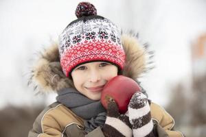 enfant dans le hiver avec magnifique fruit. une garçon dans chaud vêtements avec une rouge Pomme photo