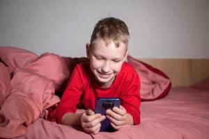 une mignonne garçon dans rouge pyjamas est mensonge dans lit en dessous de une couverture et à la recherche à le téléphone photo