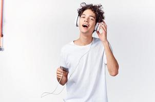 homme avec frisé cheveux dans blanc T-shirt écoute à la musique avec écouteurs téléphone La technologie photo