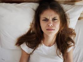 en colère femme mensonges dans lit dans une blanc T-shirt mécontent avec une du froid photo