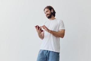 une homme avec une barbe et longue cheveux dans une blanc T-shirt et bleu jeans regards à le sien téléphone retournement par un en ligne social médias alimentation avec écouteurs dans le sien oreilles contre une blanc Contexte photo