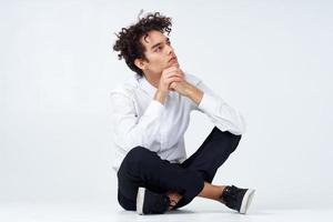 gars avec frisé cheveux dans une blanc chemise, baskets et pantalon est assis sur le sol photo