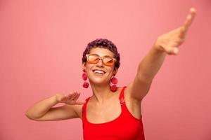 une Jeune athlétique femme avec une court la Coupe de cheveux et violet cheveux dans une rouge Haut dans des lunettes de soleil avec un athlétique figure sourit avec sa bras tendu dans différent directions dansant sur une rose Contexte photo