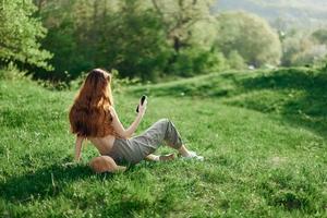 Haut vue de une femme dans un Orange Haut et vert un pantalon séance sur le été vert herbe avec sa retour à le caméra avec sa téléphone, une Jeune free-lance étudiants concept de travail et loisir photo