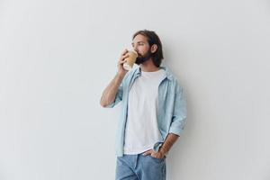 free-lance millénaire homme avec une barbe en buvant café de une recyclé tasse dans élégant branché vêtements blanc T-shirt bleu jeans et chemise sur une blanc Contexte photo