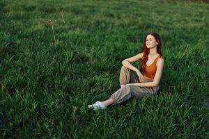 le rousse femme est assis dans le parc sur le vert herbe portant un Orange haut, vert pantalon, et baskets et regards en dehors à le réglage été Soleil photo