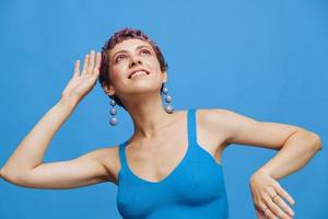 Jeune athlétique mode femme avec coloré cheveux et court la Coupe de cheveux posant et dansant dans bleu tenue de sport souriant et à la recherche à le caméra sur une bleu monochrome Contexte photo