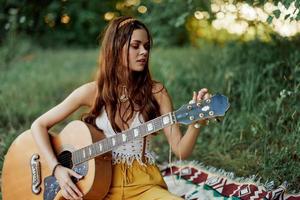 fille hippie femme en jouant guitare dans respectueux de la nature vêtements séance sur le sol à l'extérieur dans la nature dans le tomber en train de regarder le le coucher du soleil photo