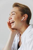 Jeune femme adolescent écoute à la musique avec infertile écouteurs et dansant maison, sourire avec les dents avec une court la Coupe de cheveux dans une blanc chemise sur une blanc Contexte. fille Naturel pose avec non filtres photo