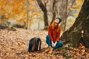 femme dans pantalon et bottes avec une sac à dos est assis près une arbre dans le l'automne forêt déchue feuilles photo