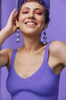 portrait de une sportif mode femme posant souriant avec les dents à le caméra avec élégant des boucles d'oreilles dans sa oreilles dans une violet yoga survêtement et une transparent casquette sur une violet monochrome Contexte photo