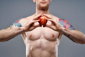 une gars avec une gonflé à bloc torse et une tatouage détient un Pomme dans le sien mains sur une gris Contexte photo