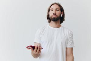 une homme avec une barbe dans blanc T-shirt et jeans regards à le sien téléphone retournement par un en ligne social médias alimentation avec écouteurs dans le sien oreilles écoute à une voix message sur une blanc Contexte et souriant photo