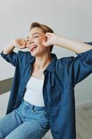 une Jeune femme séance dans une chaise à Accueil souriant avec les dents avec une court la Coupe de cheveux dans jeans et une denim chemise sur une blanc Contexte. fille Naturel pose avec non filtres photo