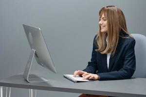 de bonne humeur millénaire blond femme d'affaires ouvrier dans bleu veste en utilisant bureau ordinateur séance à lieu de travail dans gris moderne bureau. éloigné emploi, La technologie et carrière profession concept. copie espace photo