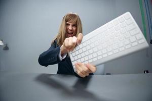 fou irrité en colère millénaire blond femme d'affaires ouvrier dans bleu veste jette clavier et en hurlant à caméra dans gris moderne bureau. gens émotions affaires concept. copie espace, large angle photo