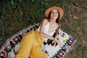 une femme dans respectueux de la nature Vêtements dans une hippie Regardez est mensonge sur une coloré plaid souriant et à la recherche à un l'automne le coucher du soleil dans la nature dans le parc. mode de vie sur une périple de harmonie avec le monde photo