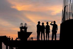 silhouette de ingénieur et ouvrier équipe vérification projet à bâtiment site arrière-plan, Infrastructure construction site à le coucher du soleil dans soir temps photo