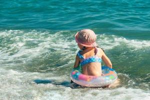 une de bonne humeur enfant d'âge préscolaire fille dans une maillot de bain et une Soleil chapeau avec un gonflable bague a amusement nager sur une ensoleillé journée dans le mer photo