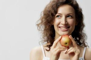 fermer portrait de de bonne humeur mignonne frisé magnifique femme dans de base blanc T-shirt prendre plaisir Pomme souriant posant isolé sur plus de blanc Contexte. Naturel respectueux de la nature des produits concept. copie espace photo