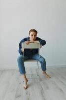 une Jeune femme séance dans une chaise à Accueil souriant avec les dents avec une court la Coupe de cheveux dans jeans et une denim chemise sur une blanc Contexte. fille Naturel pose avec non filtres photo