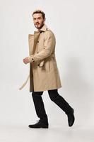une homme dans une beige manteau des promenades à le côté dans pantalon bottes sur une lumière Contexte photo