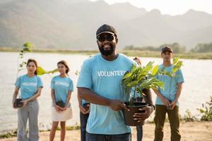 portrait de africain homme avec Contexte groupe de bénévole en portant pot avec vert plante souriant à caméra permanent sur rivière. protection de environnement et nature, écologie concept. photo