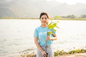 portrait de bénévole femme en portant pot avec vert plante souriant à caméra permanent sur rivière. protection de environnement et nature, écologie concept. photo