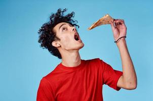 homme avec une pièce de Pizza dans le sien main sur une bleu Contexte adolescent dans une rouge T-shirt frisé cheveux photo