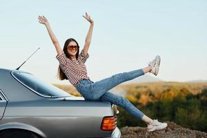 une femme avec une voiture arrêté sur le route à du repos sur le périple élevé sa bras et jambes de bonheur et une magnifique paysage photo