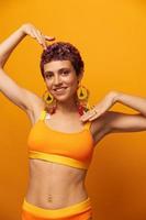 portrait de une Jeune femme avec une court la Coupe de cheveux et coloré cheveux souriant dans Jaune et montrant sa langue à le caméra sur un Orange Contexte dansant avec des boucles d'oreilles accessoires dans le studio photo